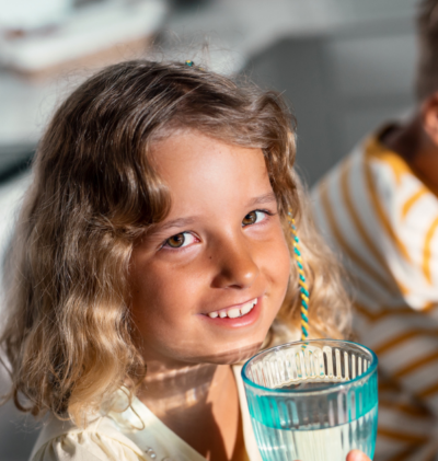 little girl at her Merced home smiles as she holds a glass of water with a little boy in the blurred background